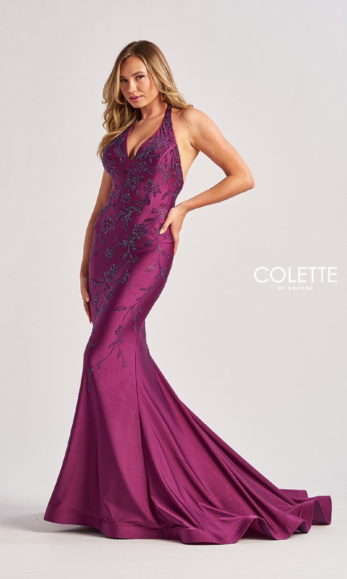 A woman poses in the Colette by Daphne Long Halter Prom Dress (CL8455), a floor-length magenta gown with floral embroidery, sleeveless design, halter v-neckline, and a flared train.